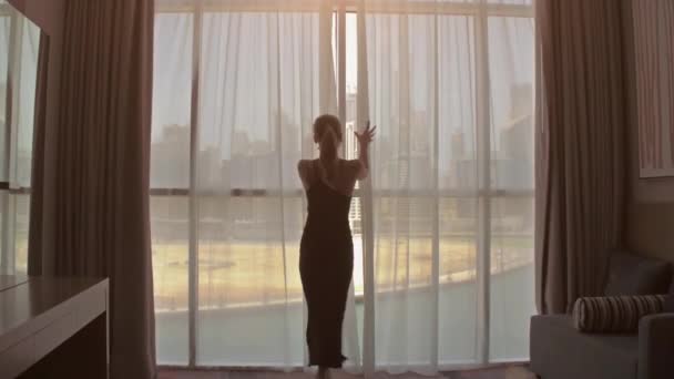 Langzame beweging. Jonge vrouw ontdekt het grote raam en kijkt uit over haar appartement op de gebouwen van de stad. Zonsopgang in de stad. — Stockvideo