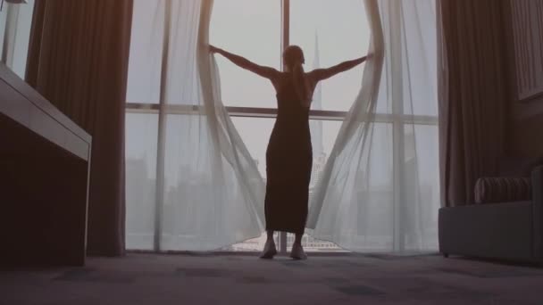 Задній вигляд жінки, що відкриває штори і дивиться у вікно, щоб прибрати в готелі вранці. Сучасні будівлі в місті Дубай. — стокове відео