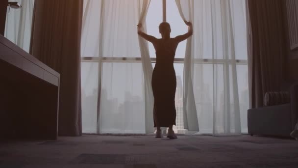 Привлекательная молодая женщина открывает занавески в замедленной съемке смотрит на восход солнца, стоящий у окна в своем доме и наслаждается видом на город с высоты — стоковое видео