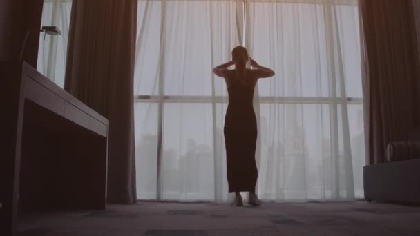 Attractive young woman opens curtains in slow motion is looking at the sunrise standing near the window in her home and enjoys of city view from height — Stock Video