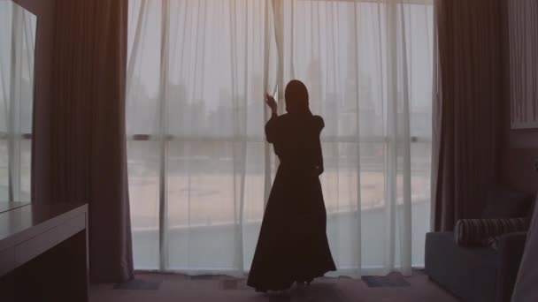 Νεαρή μουσουλμανική γυναίκα στο Ισλάμ παραδοσιακή κασκόλ κεφάλι χιτζάμπ στο σπίτι αποκαλύπτοντας το παράθυρο με θέα στην πόλη. Η πόλη του Ντουμπάι στο παρασκήνιο. — Αρχείο Βίντεο