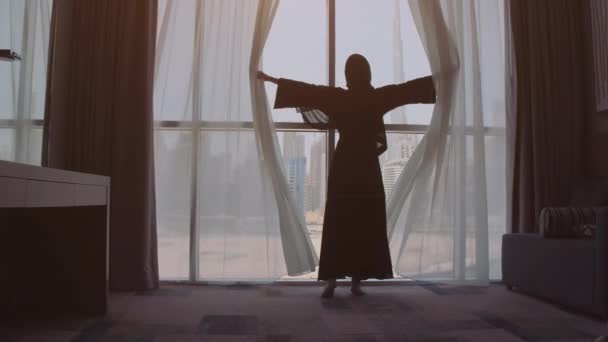 Mujer musulmana joven en el Islam tradicional pañuelo de cabeza Hijab en casa destapando ventana con vista a la ciudad. Dubai ciudad en el fondo . — Vídeos de Stock