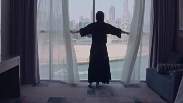 Junge muslimische Frau mit traditionellem Hijab-Kopftuch zu Hause, die Fenster mit Blick auf die Stadt freilegt. Stadt im Hintergrund. — Stockvideo