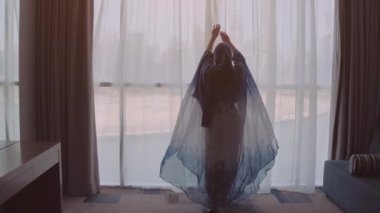 Islam 'da genç Müslüman kadın geleneksel mavi elbise evde şehir görünümü ile pencere açığa. Arka planda Dubai şehri.