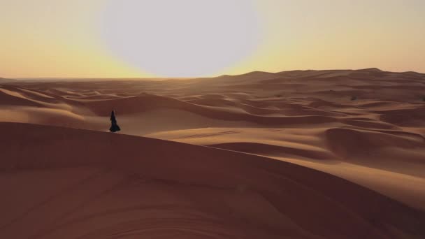 Vista aérea desde un dron volando junto a una mujer en abaya Vestido tradicional de los Emiratos Árabes Unidos caminando por las dunas en el desierto del Barrio Vacío. Abu Dhabi, Emiratos Árabes Unidos. — Vídeos de Stock