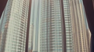 Genç kadın silueti büyük pencerenin perdelerini açıyor kollarını açıyor ve gün doğumunda apartmandaki apartmanından dışarı bakıyor. Dubai şehri