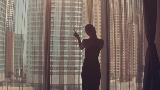 Silhueta de jovem abre cortinas na grande janela estende os braços e olhando para o seu apartamento nos edifícios da cidade durante o nascer do sol incrível. Dubai cidade — Vídeo de Stock