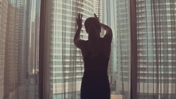 Силуэт молодой женщины открывает занавески на большом окне вытягивает руки и смотрит из квартиры на городские здания во время удивительного восхода солнца. Дубай — стоковое видео