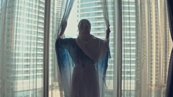 Silhueta de jovem mulher árabe vestida em vestido tradicional abaya abre cortinas na grande janela estende os braços e olhando para fora seu apartamento sobre os edifícios da cidade durante o nascer do sol incrível. Dubai cidade — Vídeo de Stock