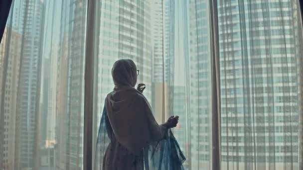 Силуэт молодой арабки в традиционной одежде Абая открывает занавески на большом окне вытягивает руки и смотрит из своей квартиры на городские здания во время удивительного восхода солнца. Дубай — стоковое видео
