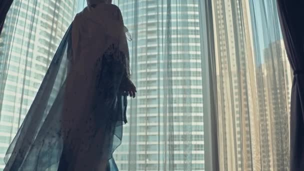Silhueta de jovem mulher árabe vestida em vestido tradicional abaya abre cortinas na grande janela estende os braços e olhando para fora seu apartamento sobre os edifícios da cidade durante o nascer do sol incrível. Dubai cidade — Vídeo de Stock