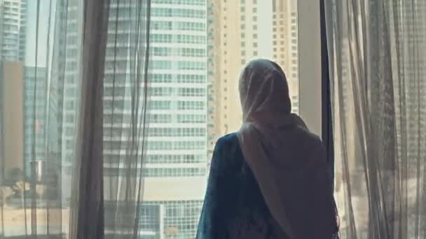 Silhouette av unga arabiska kvinnan uttröttad i traditionell klänning Abaya öppnar gardiner på det stora fönstret sträcker armar och tittar ut sin lägenhet på stadsbyggnaderna under fantastisk sol uppgång. Dubai stad — Stockvideo