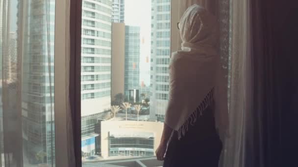 Silhouette av unga arabiska kvinnan uttröttad i traditionell klänning Abaya öppnar gardiner på det stora fönstret sträcker armar och tittar ut sin lägenhet på stadsbyggnaderna under fantastisk sol uppgång. Dubai stad — Stockvideo