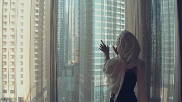전통적인 드레스 아바야에 착용 젊은 아랍 여성의 실루엣은 팔을 뻗어 놀라운 일출 동안 도시 건물에 그녀의 아파트를 찾고 큰 창에 커튼을 엽니 다. 두바이 도시 — 비디오