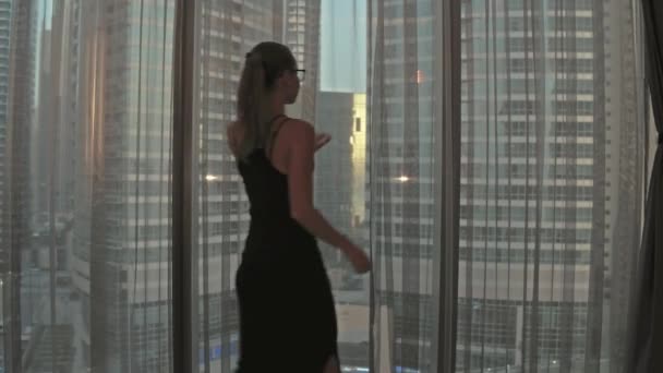 Σιλουέτα της νεαρής γυναίκας ανοίγει κουρτίνες στο μεγάλο παράθυρο απλώνει τα χέρια και κοιτάζοντας έξω από το διαμέρισμά της στα κτίρια της πόλης κατά τη διάρκεια καταπληκτική ανατολή του ηλίου. Πόλη Ντουμπάι — Αρχείο Βίντεο