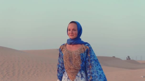 Retrato de una hermosa mujer árabe vestida con un vestido azul tradicional en el desierto al atardecer. — Vídeos de Stock