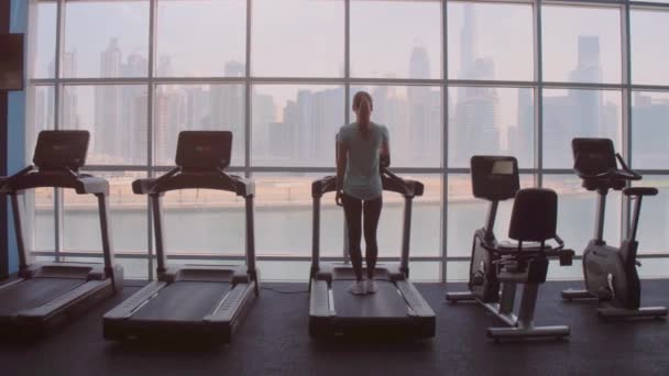 Woman running on treadmill at a panoramic window with city view at the sunrise. — Stock Video
