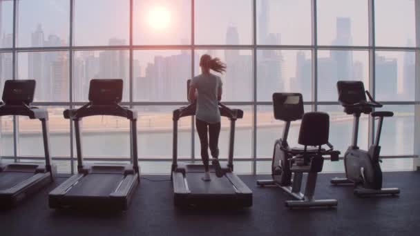 Mujer corriendo en cinta de correr en una ventana panorámica con vista a la ciudad al amanecer. — Vídeos de Stock