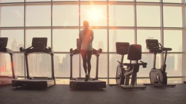 Mujer corriendo en cinta de correr en una ventana panorámica con vista a la ciudad al amanecer. — Vídeos de Stock