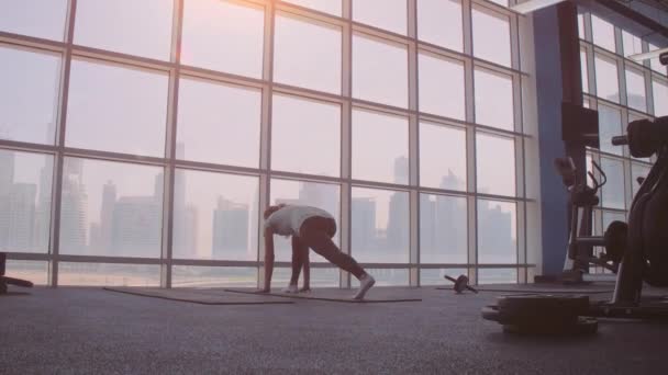 Genç bir kadın, spor salonundaki geniş panoramik pencerenin arka planında tüm vücudu minder üzerinde yoga yapıyor. Yoga eğitimi — Stok video