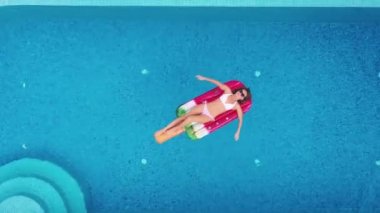 Seksi bir kadın havuz kenarında güneşleniyor ve dinleniyor. Üst kattaki havadan çekiliyor. Şişirilebilir pembe şilte görünümlü bikinili genç bir kadın.