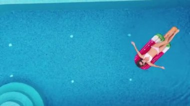 Seksi bir kadın havuz kenarında güneşleniyor ve dinleniyor. Üst kattaki havadan çekiliyor. Şişirilebilir pembe şilte görünümlü bikinili genç bir kadın.