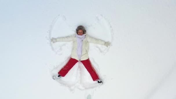 Uma jovem deitada na neve está pintando um anjo. A câmera gira em torno de e zooms em cima dela. Férias de Natal. — Vídeo de Stock