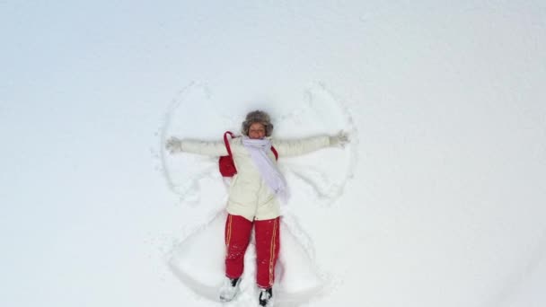 Uma jovem deitada na neve está pintando um anjo. A câmera gira em torno de e zooms em cima dela. Férias de Natal. — Vídeo de Stock