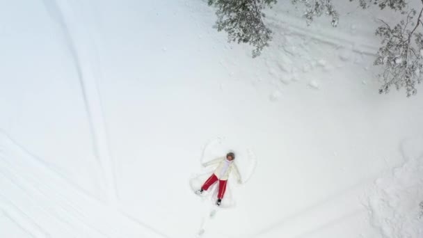 航空機だ。雪の中に寝そべっている少女が天使を描いている。カメラは周りを回り、彼女の上にズームします。クリスマス休暇. — ストック動画