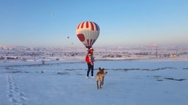 Kışlık kıyafetler giymiş genç bir kadın sıcak hava balonlarına bakıyor ve köpeğiyle oynuyor. İnanılmaz seyahat konsepti. 