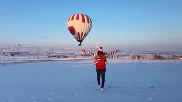 Młoda dziewczyna idzie przez pole zimowe, widzi wielokolorowy balon na gorące powietrze i wznosi ramiona patrząc na piękny krajobraz. — Wideo stockowe