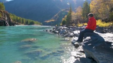 Dağ manzaralı bir nehrin yanında bir kadın oturur