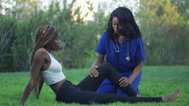 慢动作射击在一个黑人妇女在一个公园在磨砂帮助另一个妇女与她的腿 而坐在草丛中 — 图库视频影像