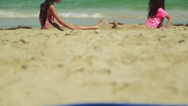 Tilt up of two young girls sitting by the ocean. One of them lays down and starts making sand angels.