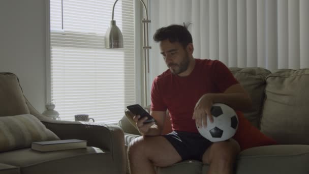 Hombre en el sofá sosteniendo la pelota de fútbol y pasando por juega en su teléfono celular — Vídeos de Stock