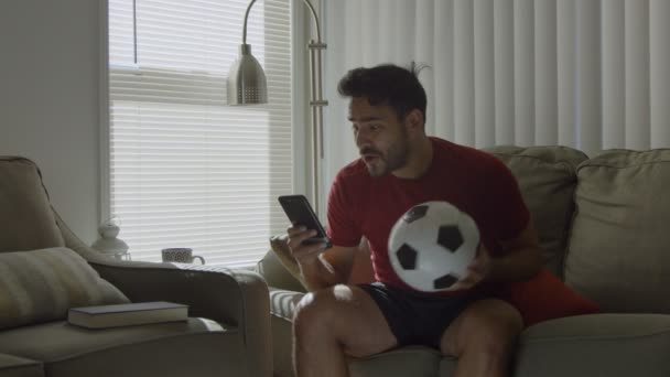 Hombre feliz en casa viendo el partido de fútbol en el teléfono celular mientras sostiene la pelota de fútbol — Vídeos de Stock