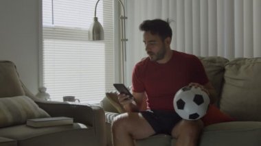 Evde futbol topu tutarken cep telefonundan futbol maçı izleyen adam