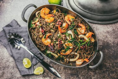 Buğday noodle ile deniz ürünleri - karides, ahtapot, kalamar metal tencerede. Üstten görünüm, taş arka plan. Tonda.