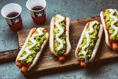 Chilean Completo Italiano. Hot dog sandwiches with tomato, avocado and mayonnaise served on wooden board with drink in paper cup . Top view. Independence Day concept. clipart