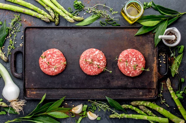 Raw meet ground beef burgers on cast iron frying board with asparagus, herb, oil and spices. Top view.