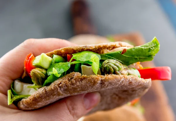 Hand holding vegetarian pita sandwich stuffed with asparagus, bell pepper, avocado and fresh leaves.