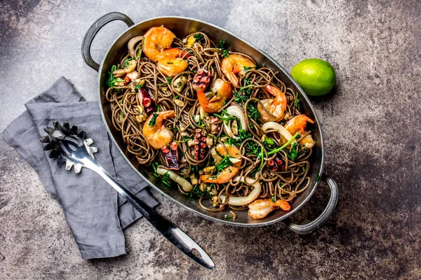 Buğday noodle ile deniz ürünleri - karides, ahtapot, kalamar metal tencerede. Üstten görünüm, taş arka plan.