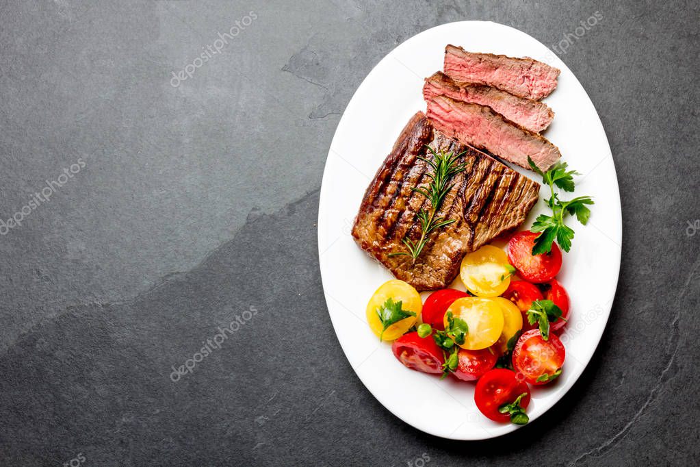 Sliced medium rare grilled beef steak served on white plate with tomato salad and potatoes balls. Barbecue, bbq meat beef tenderloin. Top view, slate background, copy space.