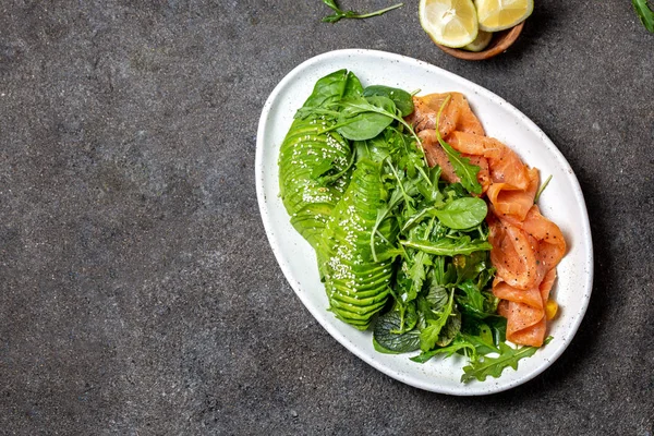 Low carbs salad. Spinach, rucola salad with avocado and salmon. Black concrete background, white plate, top view — Stock Photo, Image