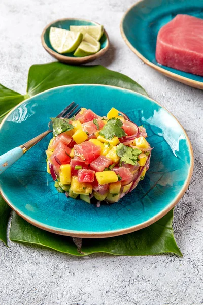 Tártaro de ensalada de mango de atún gourmet de estilo tropical con cilantro y cebolla morada. En plato azul, hojas tropicales — Foto de Stock