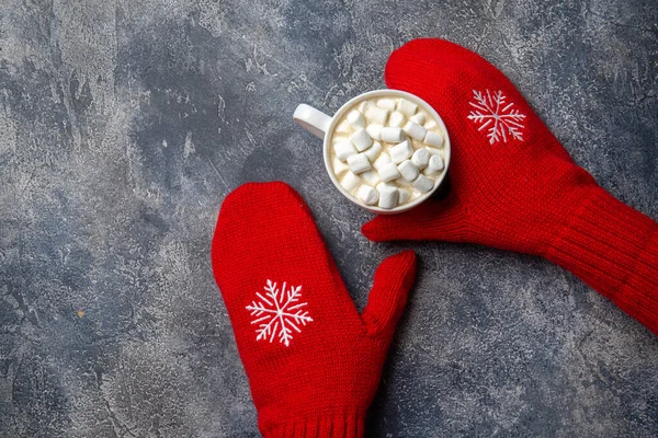 Natal e Ano Novo composição de férias acolhedora com cachecol, mãos de mulher em mitenes, canecas com bebida quente e marshmallow no fundo de concreto cinza. Deitado plano, vista superior. — Fotografia de Stock