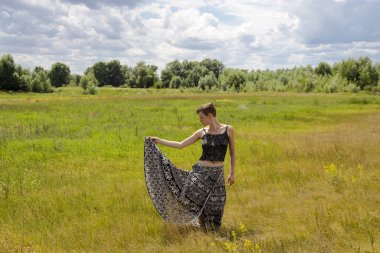 Siyah-beyaz etno desenli uzun etekli kız, tek eliyle eteğini kaldırıyor, sarı çiçekli bir tarlanın arka planına doğru.