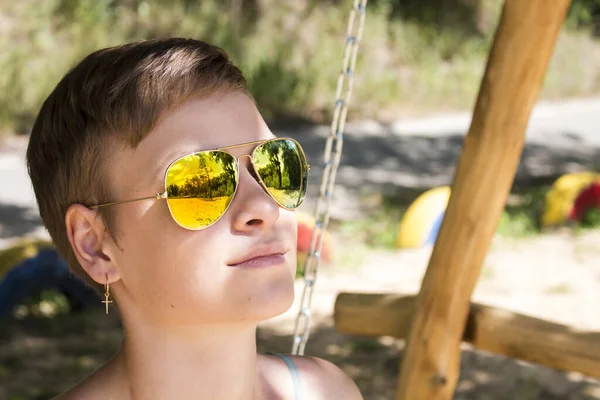 Portrait of a short-cut girl in yellow sunglasses, in whose lenses trees are reflected