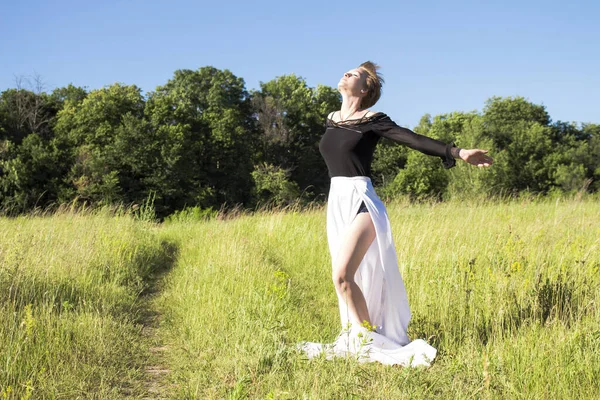 A short-cropped girl in a black blouse and a long white skirt, puts her face to the sun\'s rays and the wind, her arms outstretched against the background of nature