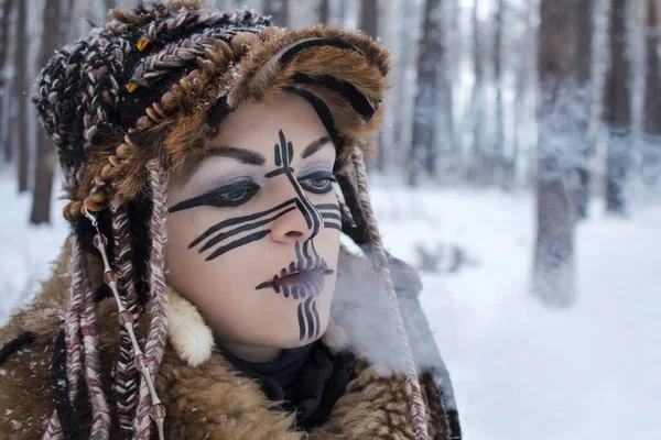 A girl dressed as a wild animal hunter from animal fur, exhales steam from her mouth in front of her, against the background of a winter forest covered with snow